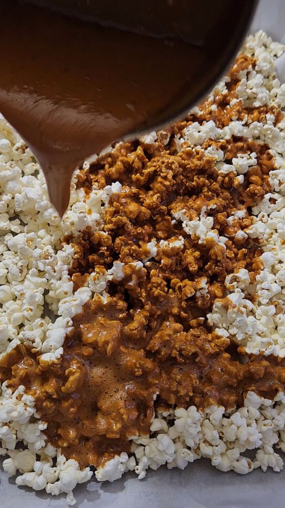 Pouring sugar-free caramel sauce onto plain popcorn for low-carb caramel popcorn made without corn syrup.