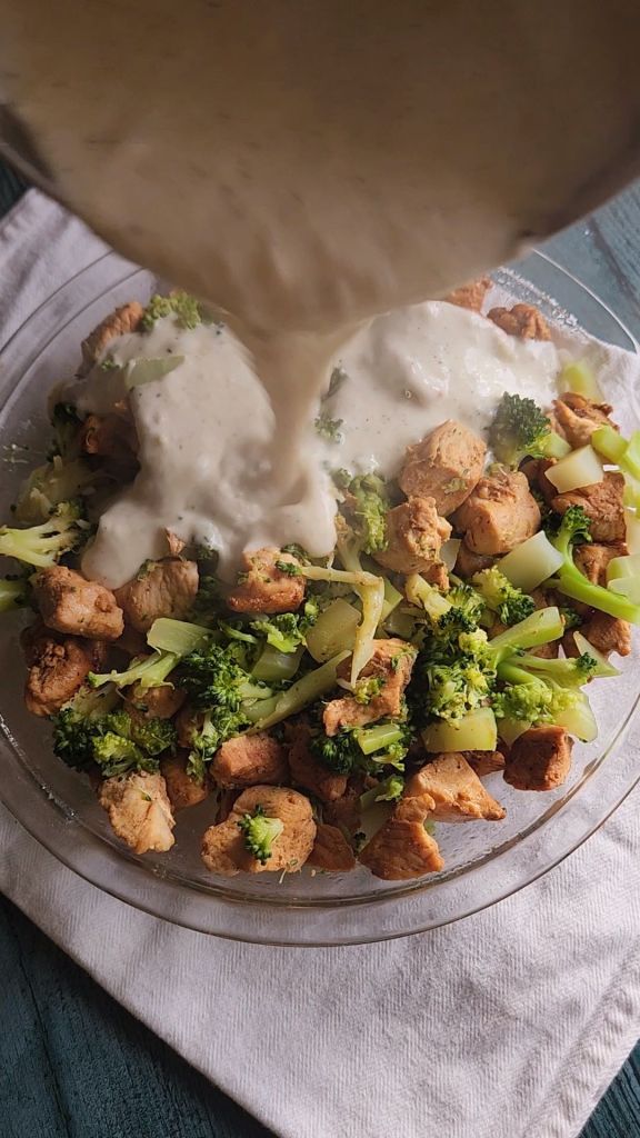 Pouring homemade cream soup over chicken and broccoli for Easy gluten-free chicken Divan casserole.