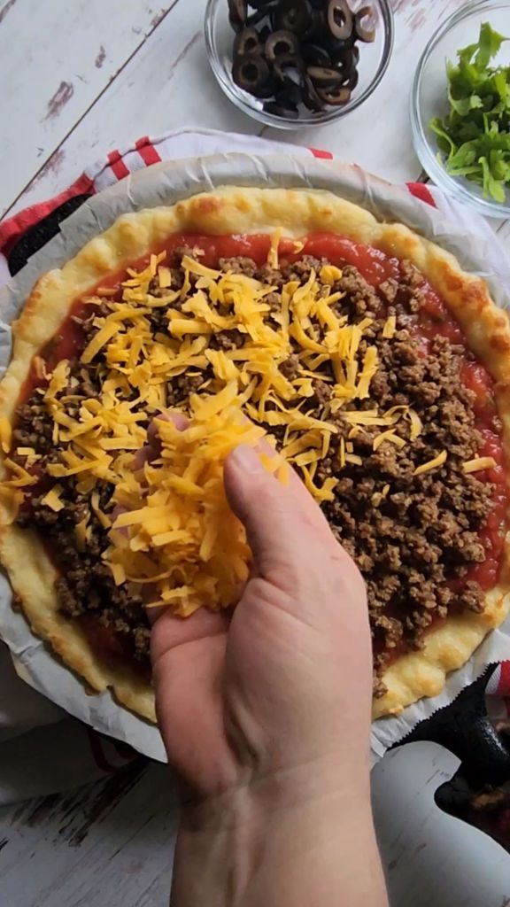 Sprinkling cheddar cheese over taco meat layer for gluten-free copycat Godfather's taco pizza.