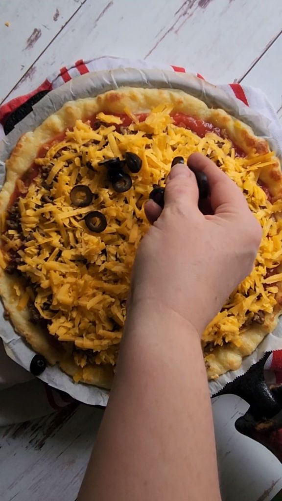 Sprinkling black olives over cheddar cheese layer for gluten-free copycat Godfather's taco pizza.