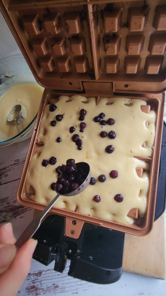 Sprinkling blueberries over Easy Gluten-Free Dairy-Free Blueberry Waffle batter in waffleiron.